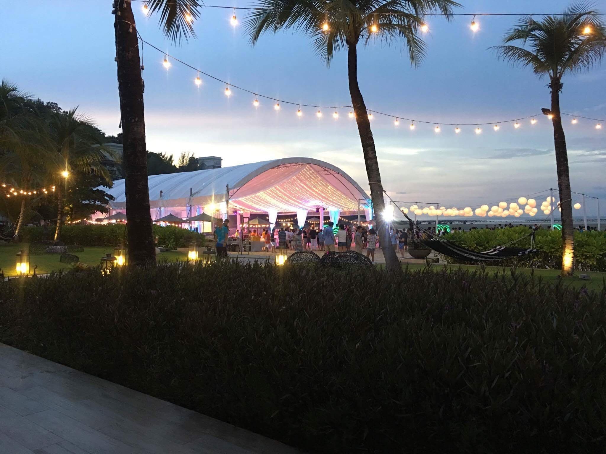 The retreat's illuminated marquee and guests beside the beach at dusk, beneath palm trees and strings of lights.